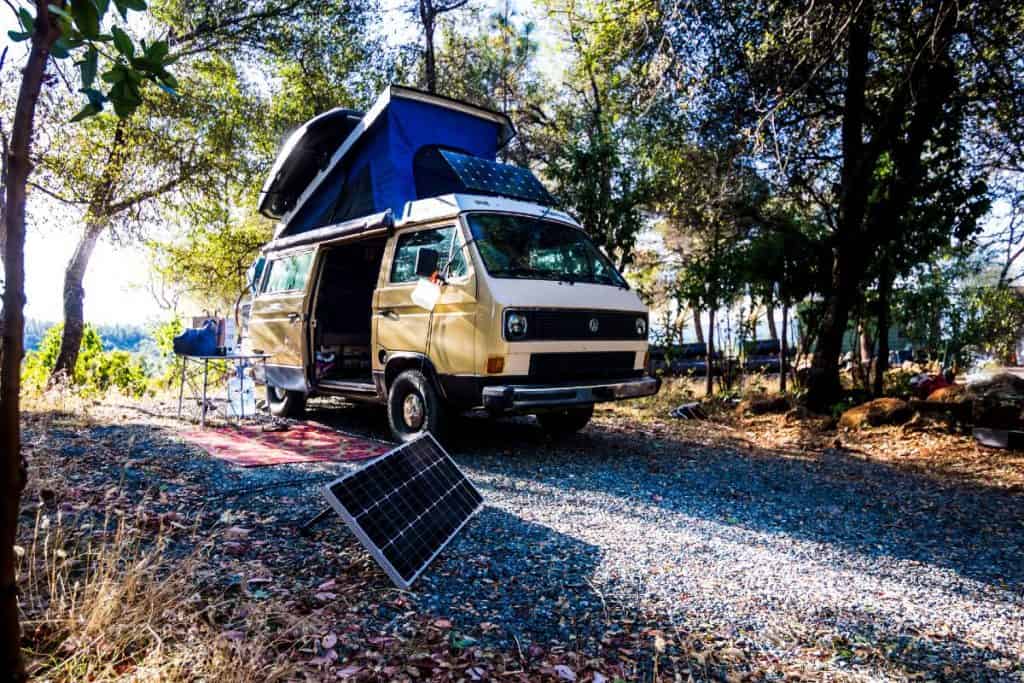 The van is parked under trees with the solar panels propped up.