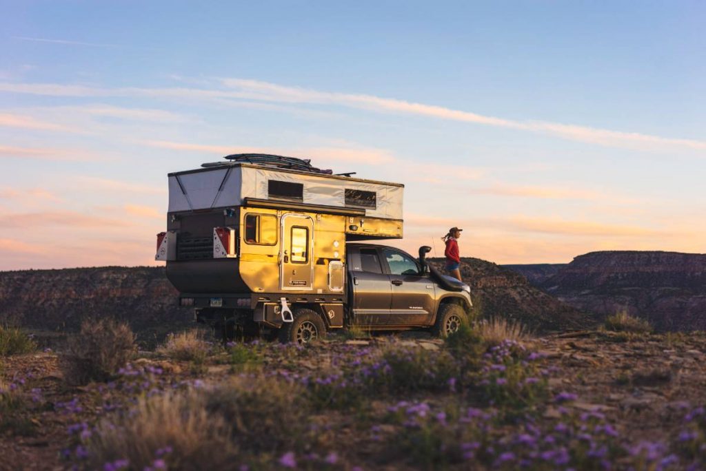 Toyota truck camper garé sur le rebord d'un canyon, avec une femme debout sur le pare-chocs avant qui regarde à l'extérieur