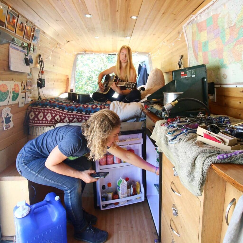 Two women inside a converted sprinter van. One sits on the bed playing the guitar, while the other grabs something out of the fridge