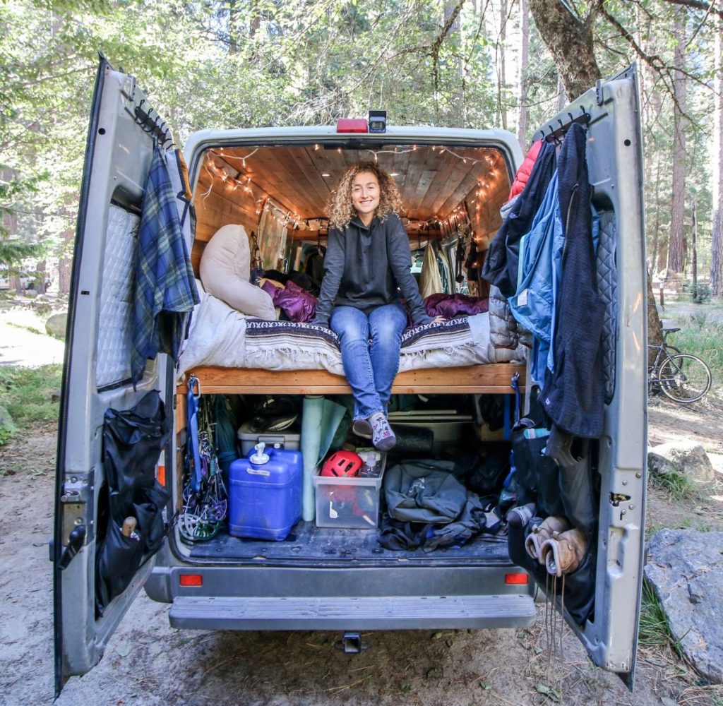 Showing inside the back door of a sprinter van, with a woman sitting on the bed