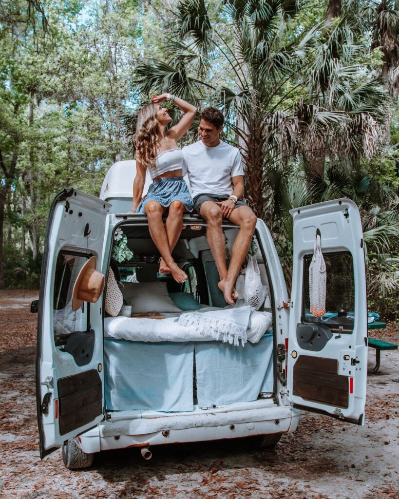@theotherside_vlog Couple sitting on top of a white ford transit connect camper van with open rear door