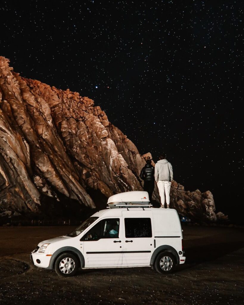 theotherside_vlog White transit connect camper van parked near boulders with two people looking at the stars