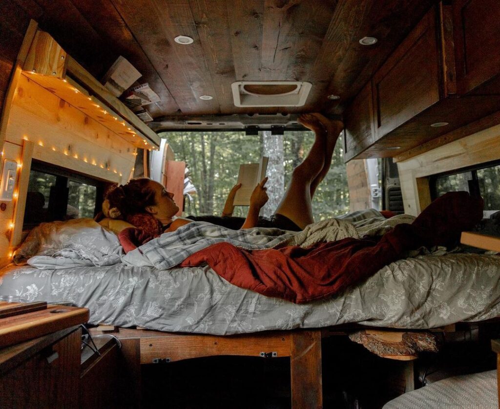 @linnea.and.akela Woman reading a book on bed in her ram promaster camper van