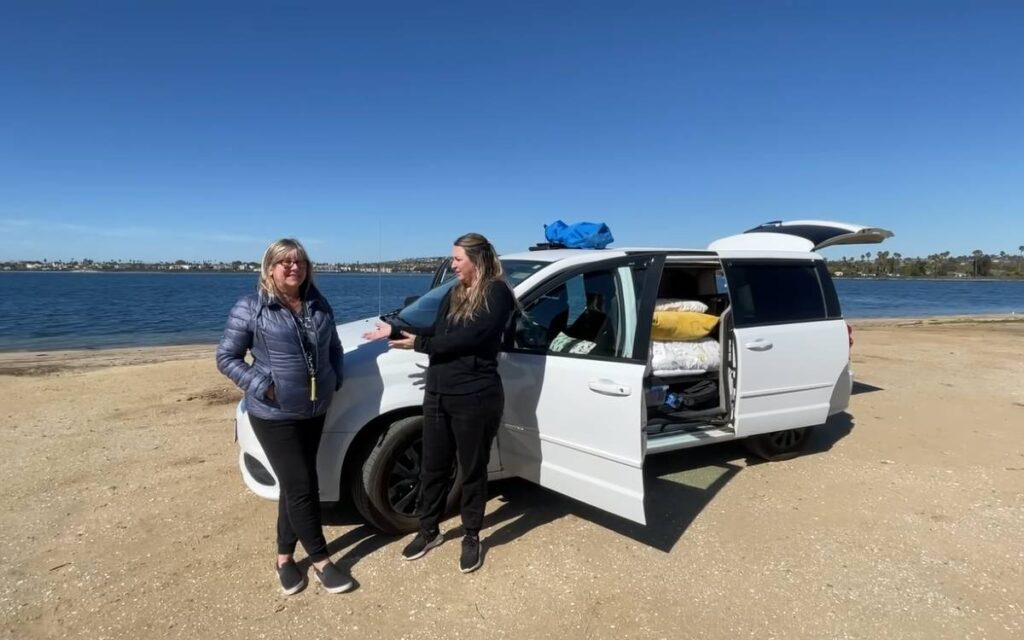 Adriana with her friend standing next to a mini van camper parked on a beach