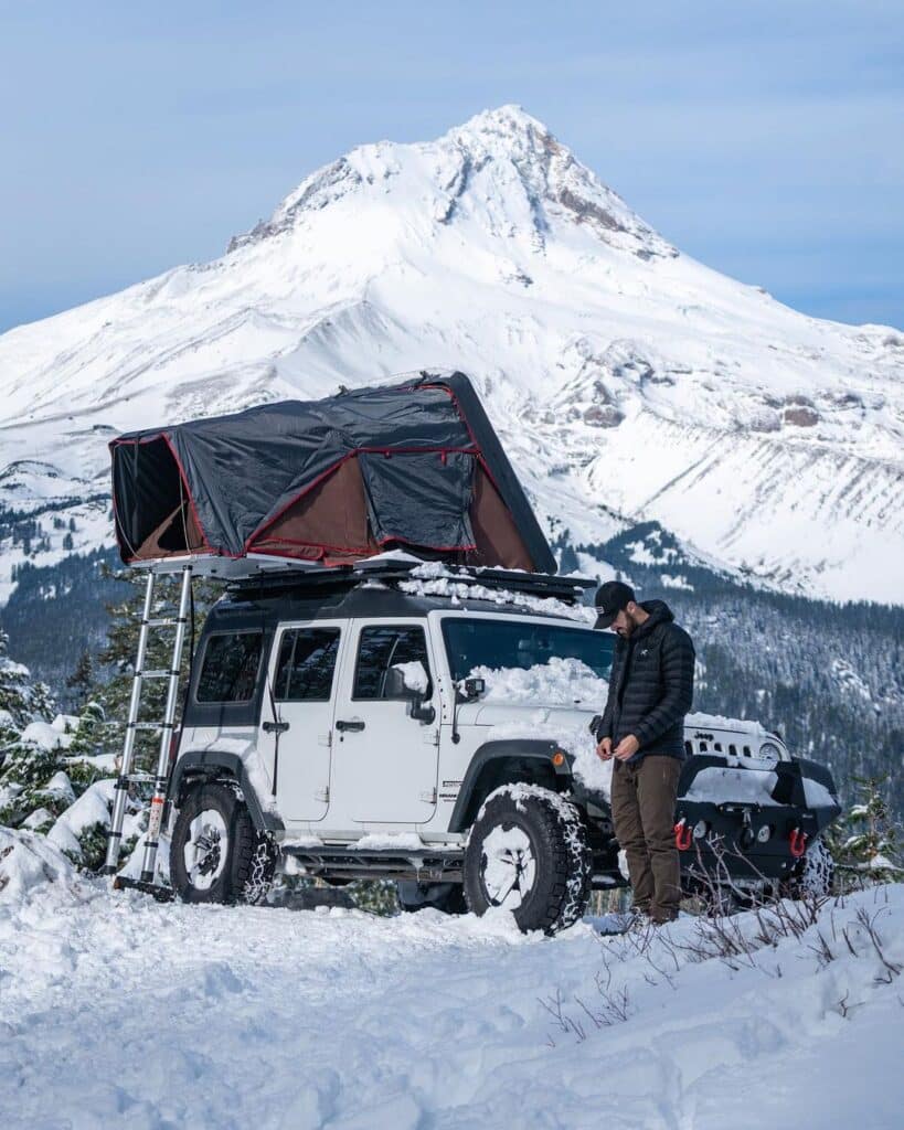 @drew.simms Man standing next to an suv camper with beautiful snowy mountain view on the background