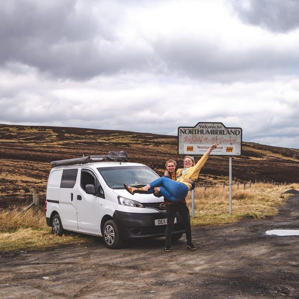 @justuswandering Man carrying a woman in his arms in front of nissan nv200 camper van