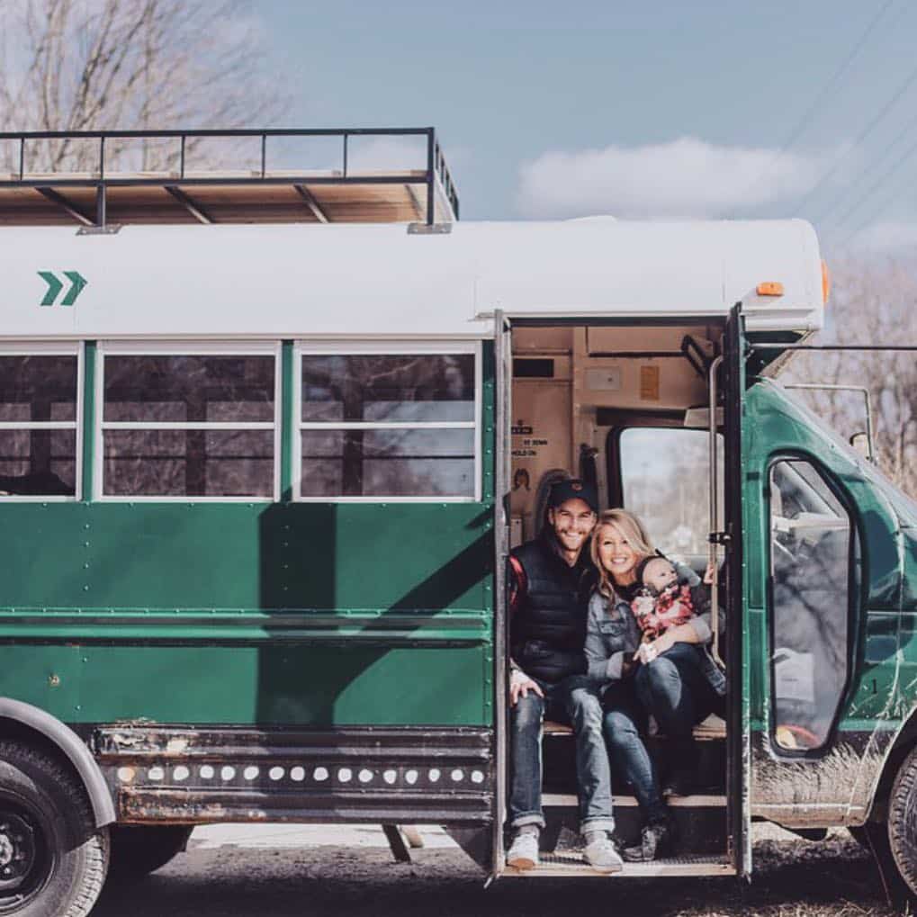 @greenbusandus Young couple with a baby sitting at the door of a short bus converted to camper