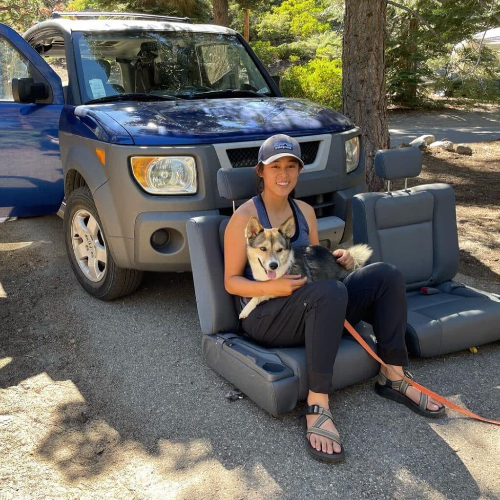 @amyismakingstuff Amy sitting with her dog on her lap in one of the two car seats placed on the ground, in front of her honda element RV camper