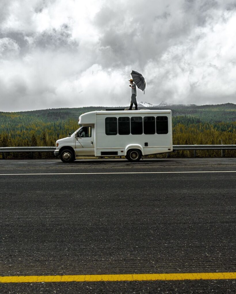 @nomad_brad_503 Brad holding an umbrella while standing on the roof of his minibus camper, parked on the side of a road