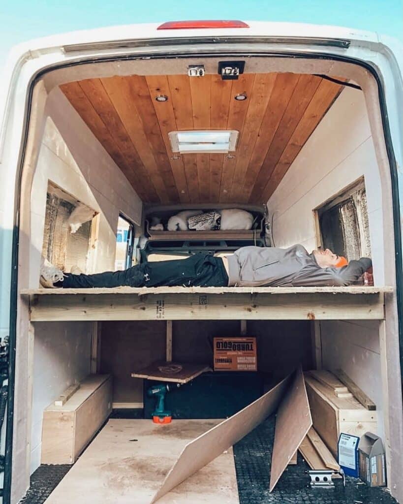 Man lying on his back inside an unfinished camper van, thinking of camper van bed ideas