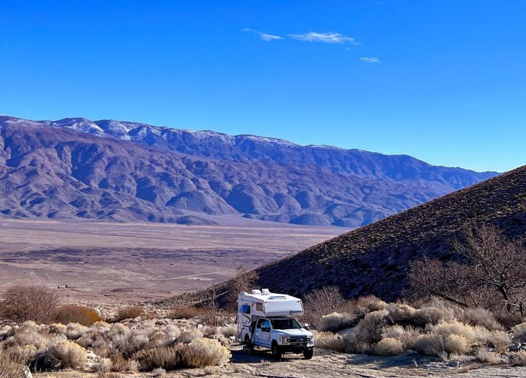 White campervan on a dirt road, overlooking a desert field with mountains under blue sky