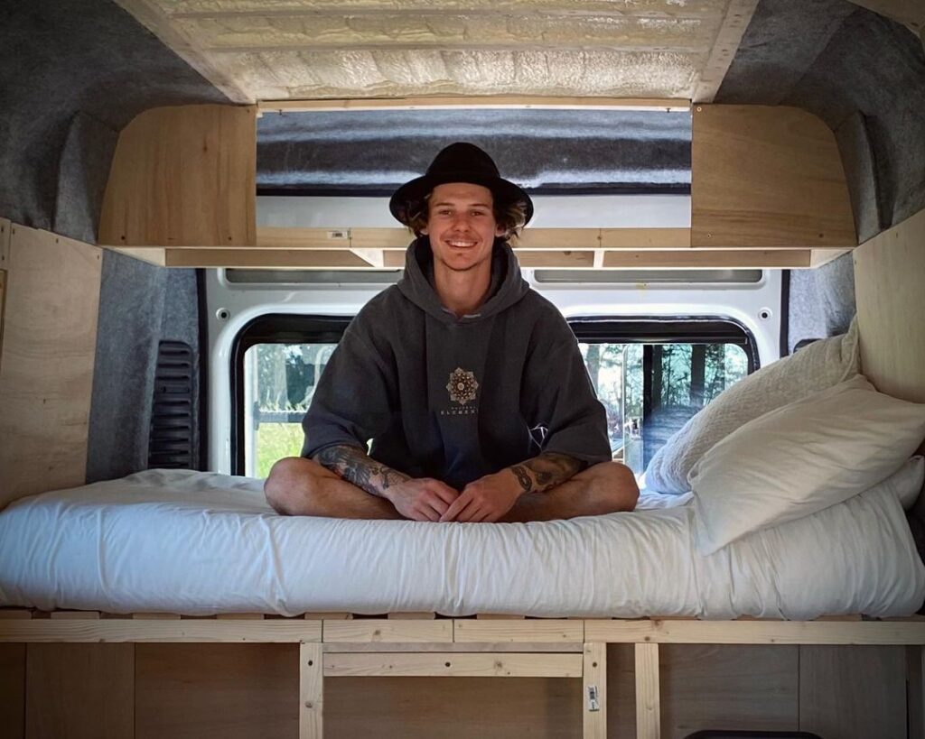 Young man wearing a hat sitting cross legged on his campervan bed