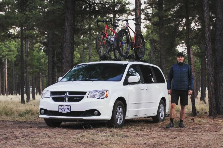 @bicycletouringpro Darren standing in the woods next to his white dodge grand caravan camper with two bicycles on roof racks