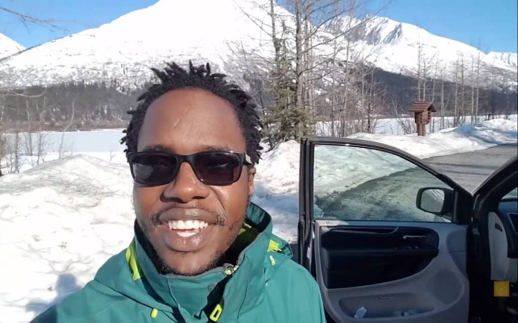 @divinezeal Nobi filming himself outside his Grand Caravan parked near a frozen lake with snow-capped mountain in the distance
