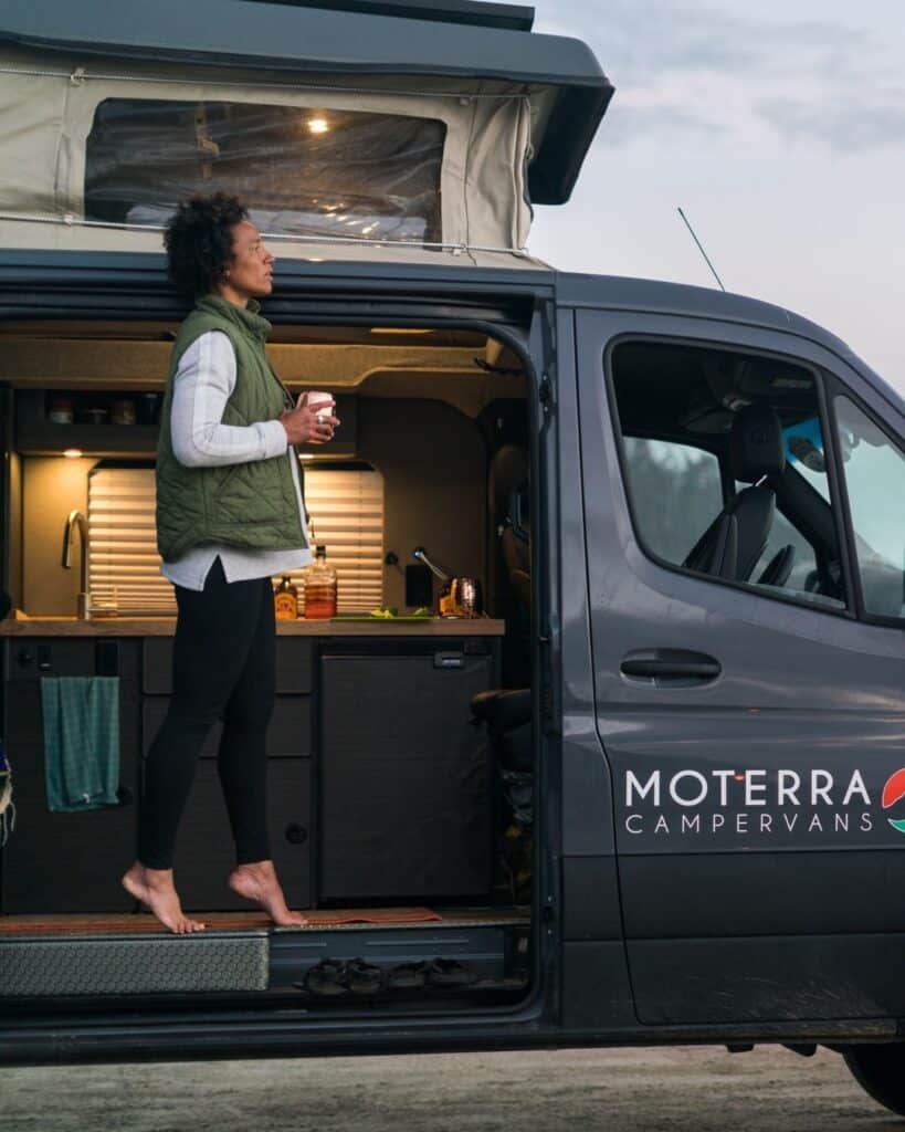 @moterra Side view of a woman holding a white coffee cup while standing on tiptoes at camper van rentals door