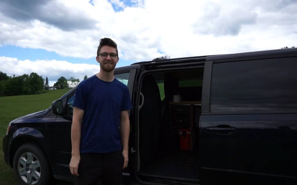 @nickandmaela Nick smiling and looking at the camera while standing next to his black dodge grand caravan camper