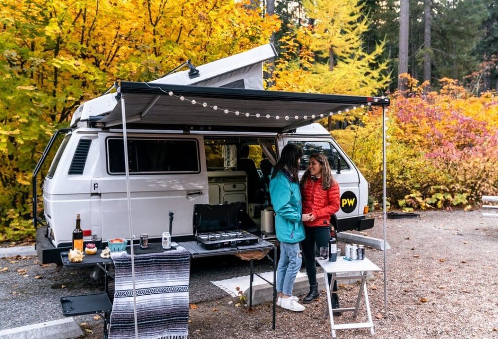 @pacwesty two women holding hands in front of a vw vanagon parked in the woods with awning extended, tables and chairs set up underneath