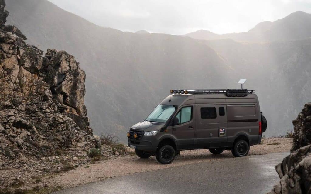 @the4x4spaceship Gray sprinter campervan parked on a cliff against beautiful mountain and blue sky background