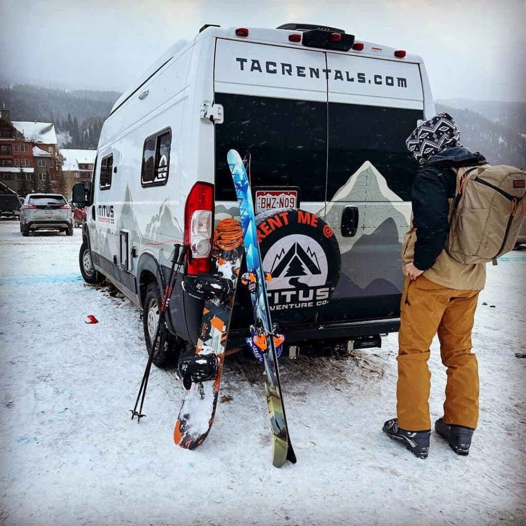 @titusadventureco Man with ski gear standing next to a campervan rentals vehicle for winter camping