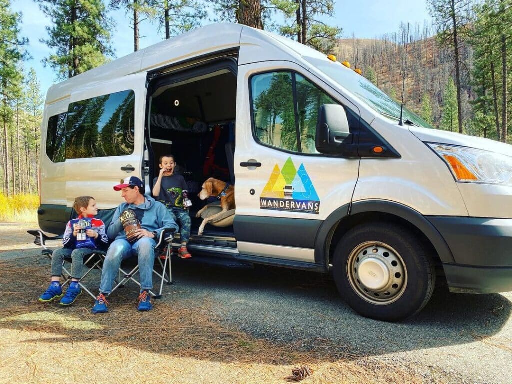 @wandervans Man with his two sons and dog eating snacks, enjoying camping in the forest in a campervan rentals camper