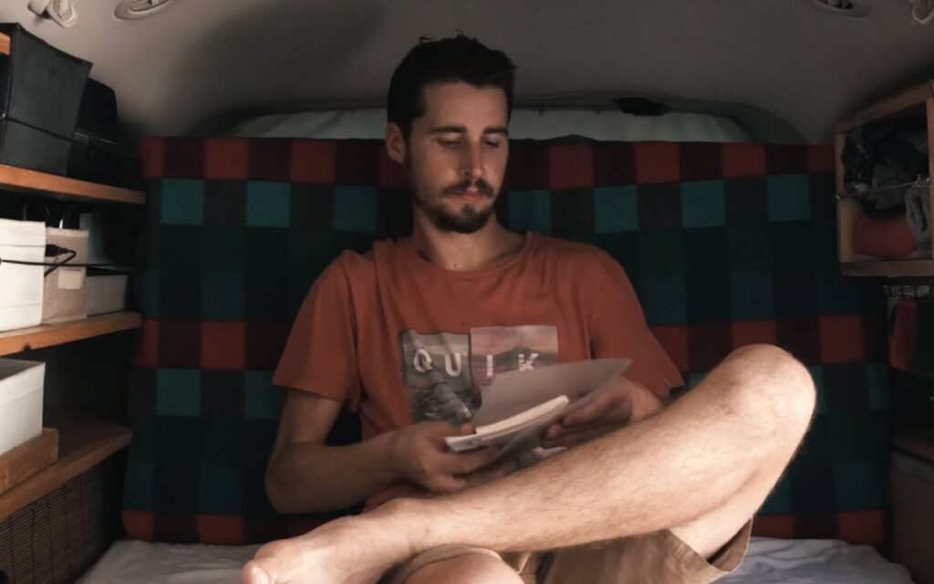 @whatsup.onearth Dodge grand caravan minivan camper conversion interior, Elian sitting cross legged on a sofa bed while looking down at the papers on his hands