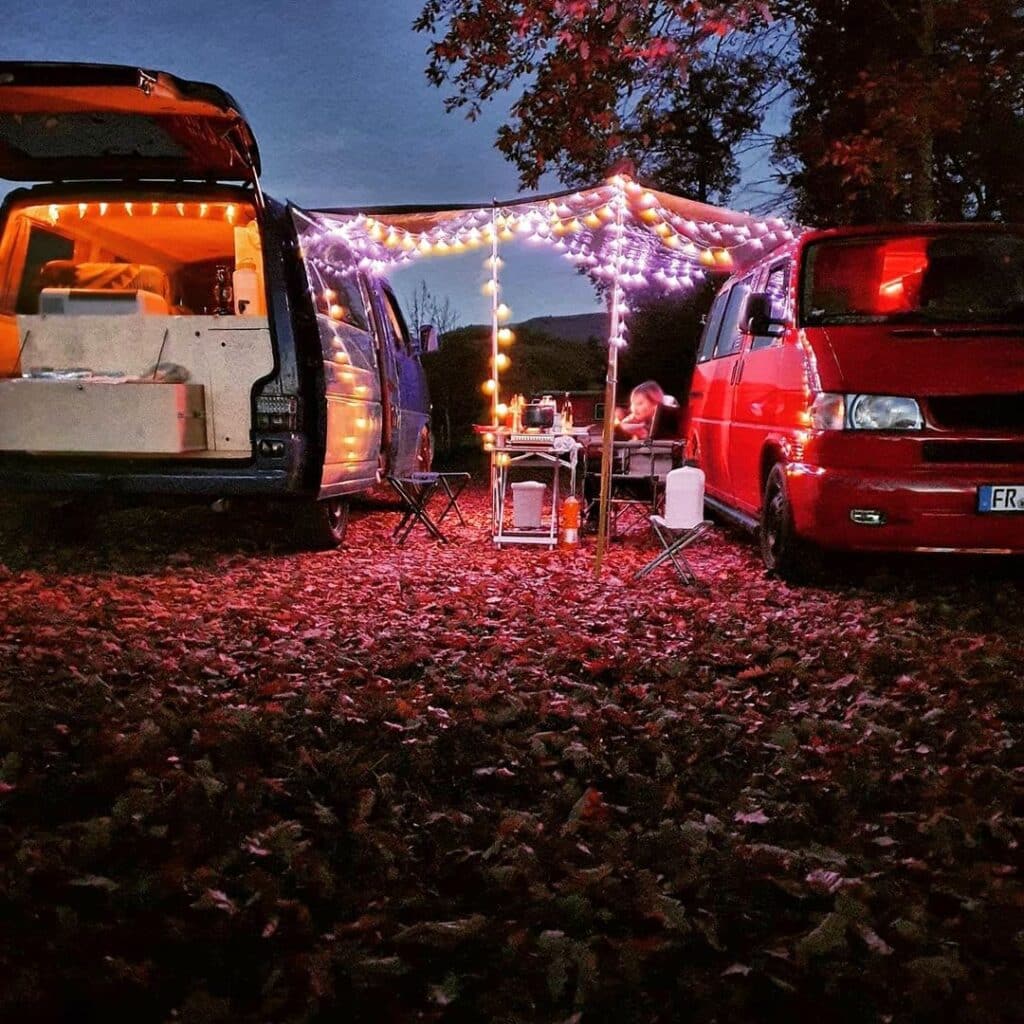 @lady_red_bulli Woman enjoying a cozy night sitting outside two campervans decorated with fairy lights