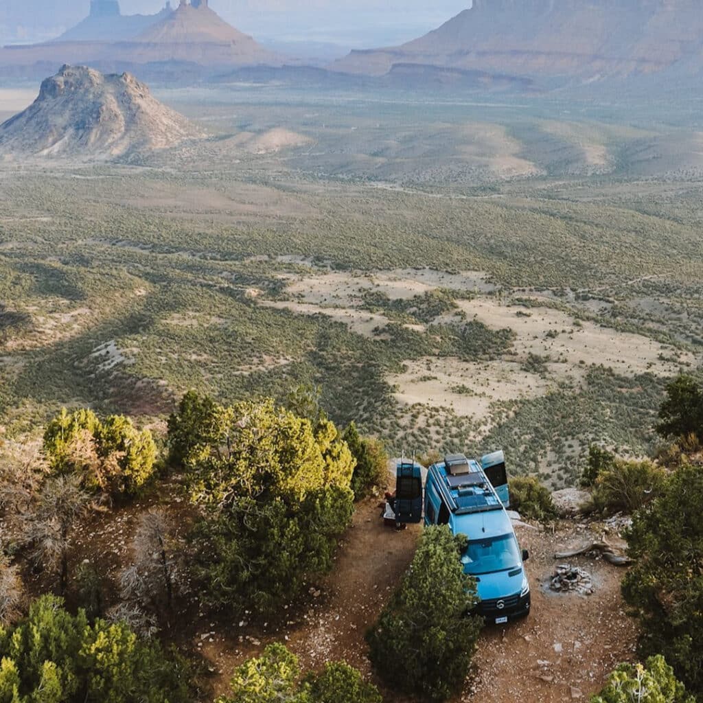 @myrecvan Aerial shot of a blue campervan parked near the edge of a cliff overlooking the mountains and forest