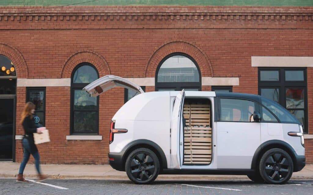 Woman holding a box while walking towards a white Canoo LDV van parked in front of a brick building