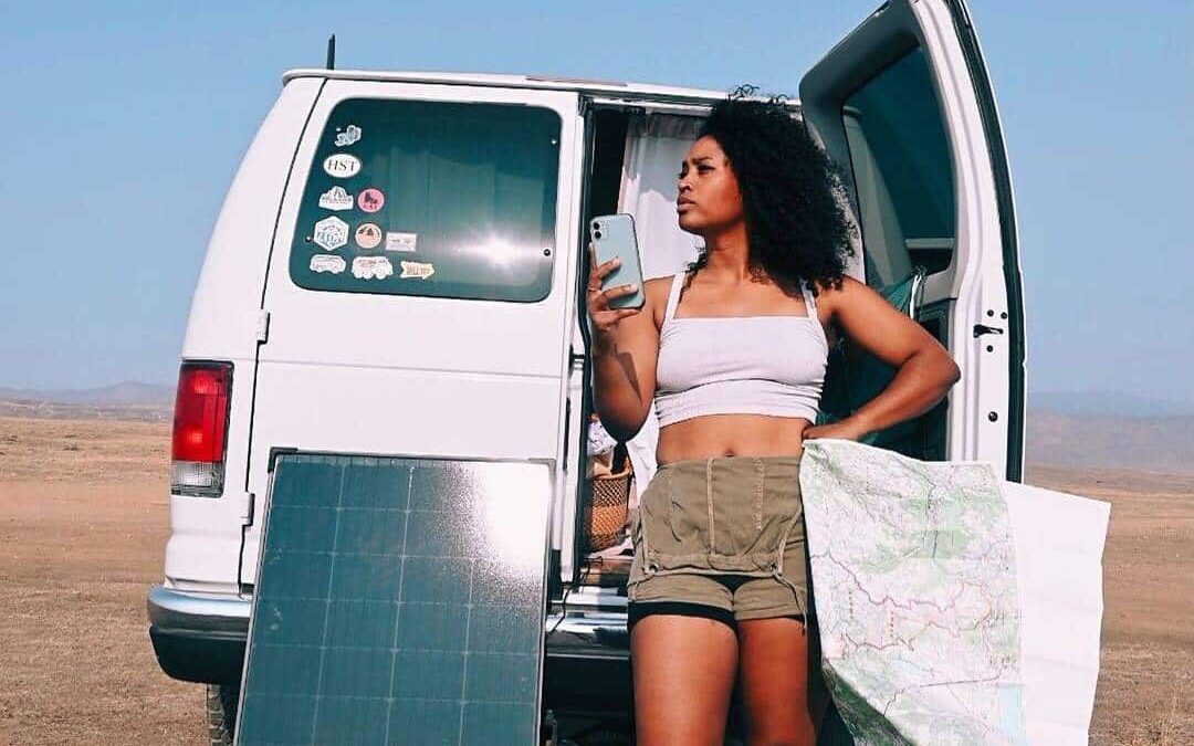 @tashofalltrades Solar panel leaning against the back of a white camper van, a woman is next to it posing at the camera while holding her smartphone