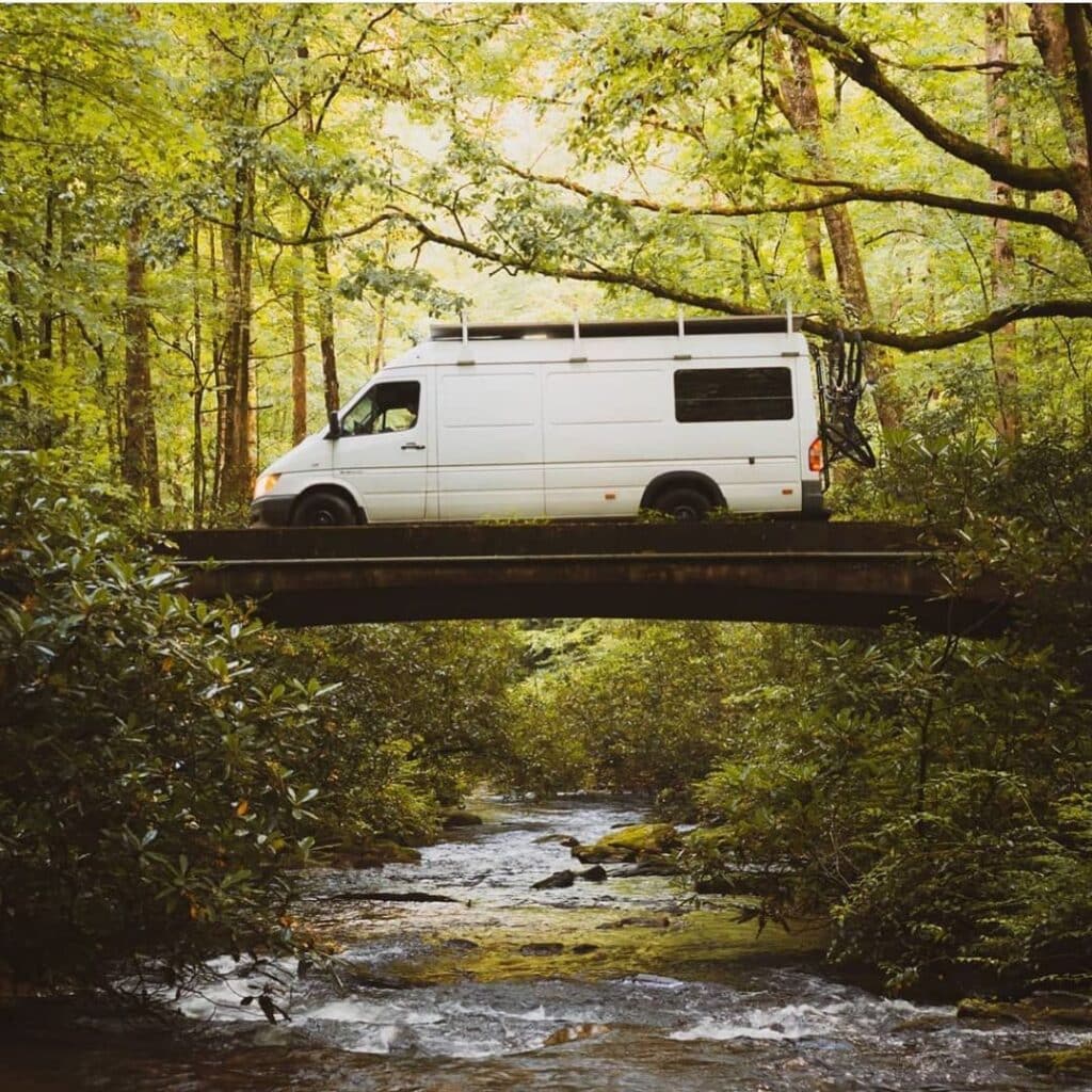 @eastcoast.vanlife Side view of a white camper van on a small bridge in the middle of a forest