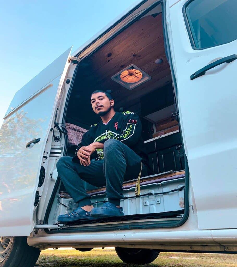 @goldenboyvanlife Ground view of a man sitting at his campervan door looking down at the camera