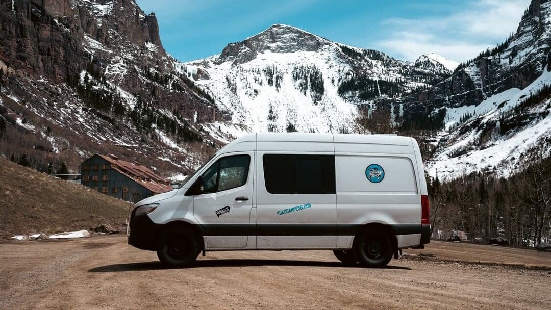 @kukucampersusa white Sprinter van parked on dirty road with snow-capped mountain view in the background