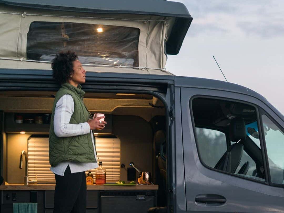 @moterra Side view of a woman holding a white coffee cup while standing at the side door of a van rental from moterra campervans
