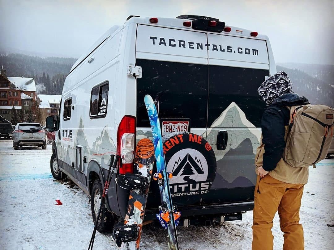 @titusadventureco Man with ski gear standing behind a rented van in a snowy mountain town