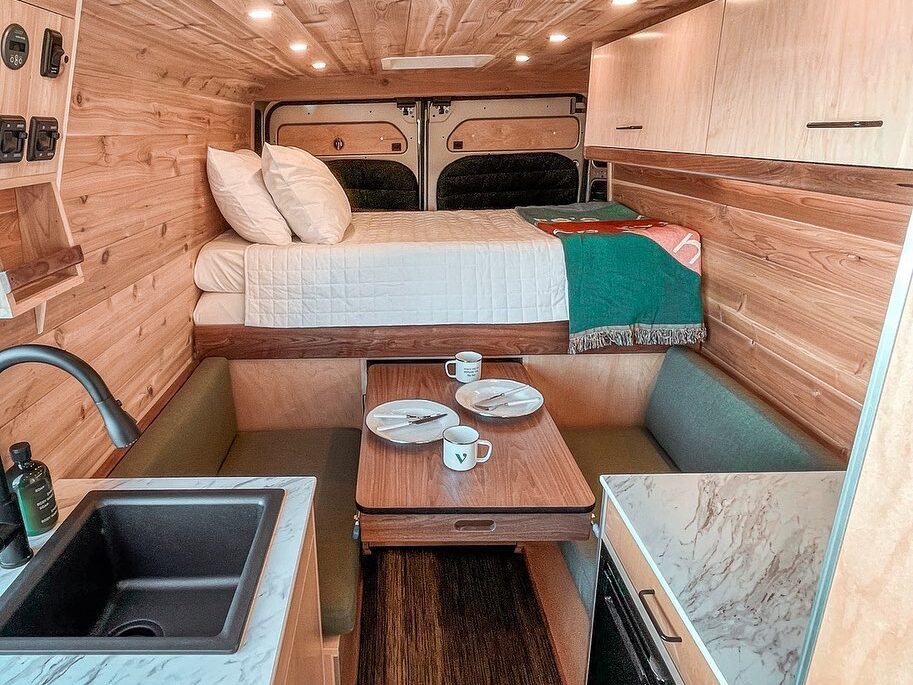 @vanishtravelco interior of a van with beautiful wood paneling, dinette, sink, fridge, and bed across the back